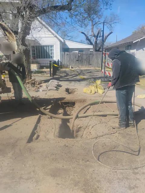 Two workers using a vacuum hose and a water wand to dig a hole near the street in front of a house, with barricades and caution tape