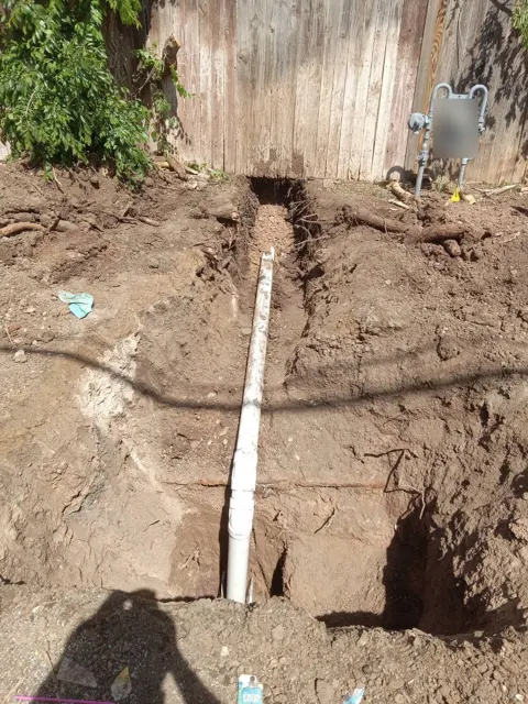 A dug-out trench in a backyard with a long run of white PVC drain pipe exposed at the bottom, next to a wooden fence