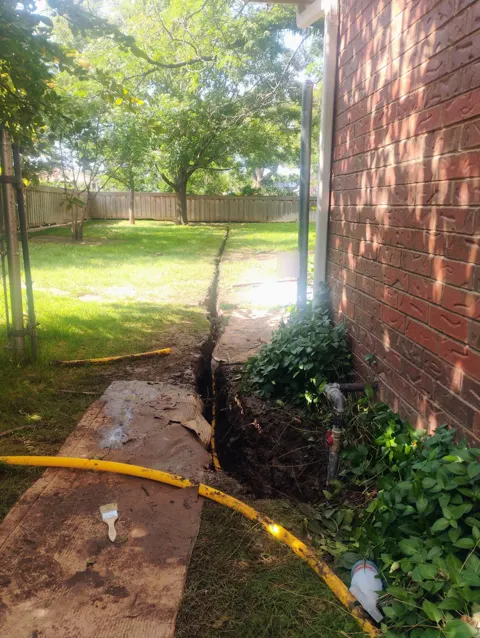 A trench dug along the side of a brick house, with yellow line laid on the ground and an outdoor faucet valve on the wall