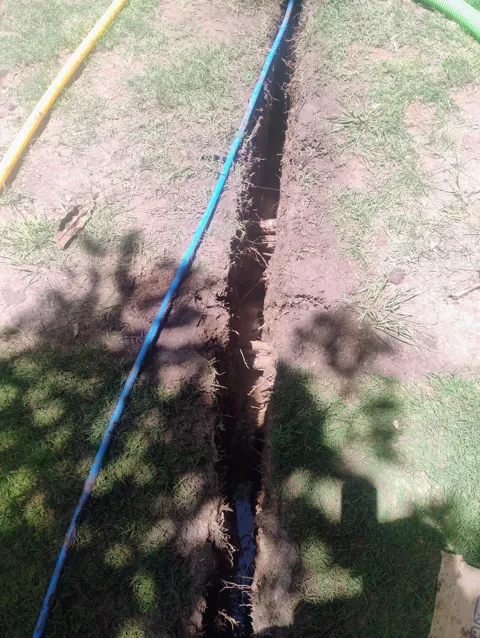 A narrow trench cut through a lawn with a blue plastic water service pipe laid on the grass beside it