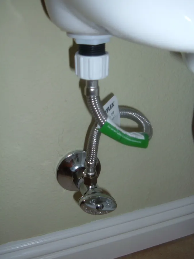 Chrome shutoff valve on the wall behind a toilet with a braided stainless supply line looping up to the fill valve